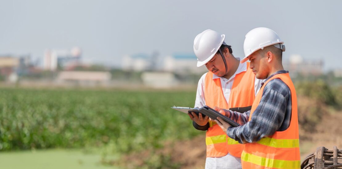 Two environmental professionals in hard hats and safety vests review site information beside a green waterway.