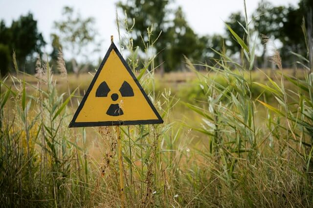 Yellow triangular radiation warning sign surrounded by tall grass, with trees in the background. radon.