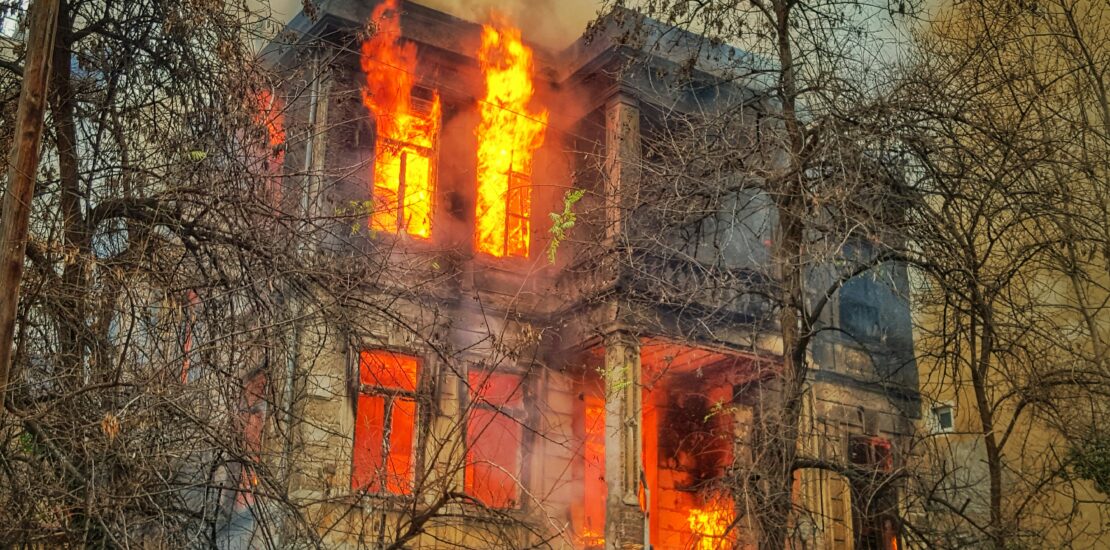 Flames and smoke pouring from the windows of a two-storey building, partially obscured by bare tree branches.